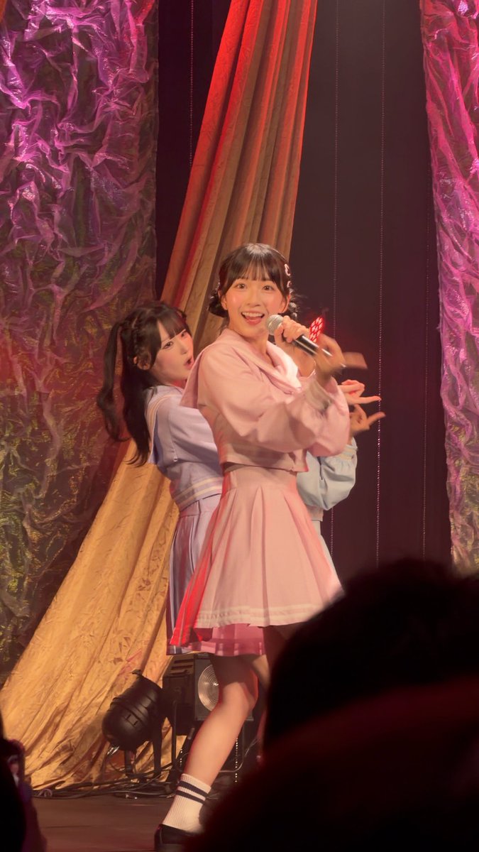 Two female performers in pink and blue outfits perform on stage, with one holding a microphone.