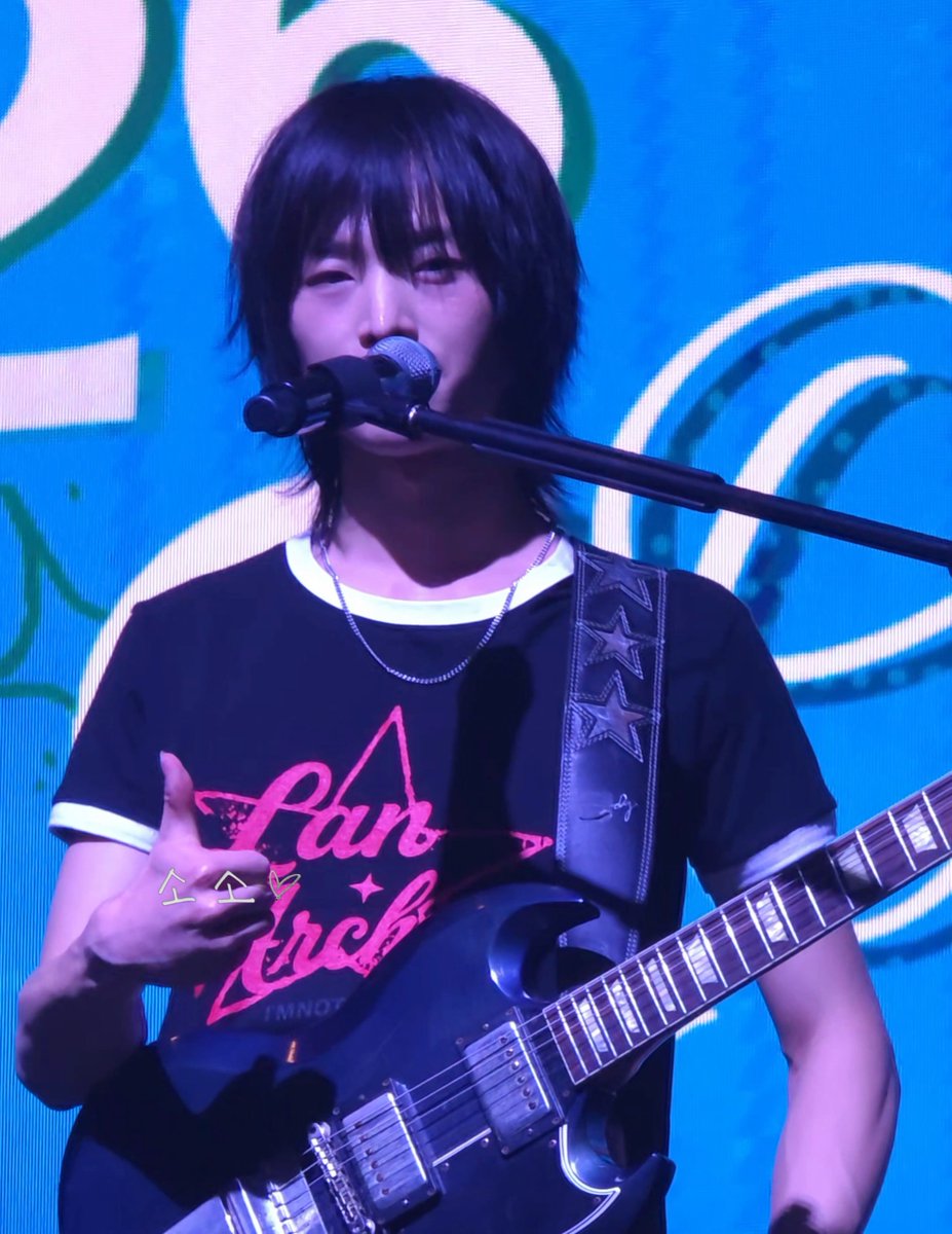A musician with dark hair plays a black electric guitar and sings into a microphone on a stage with a blue background, wearing a black t-shirt with a pink star and 'Pan* Arch' text.