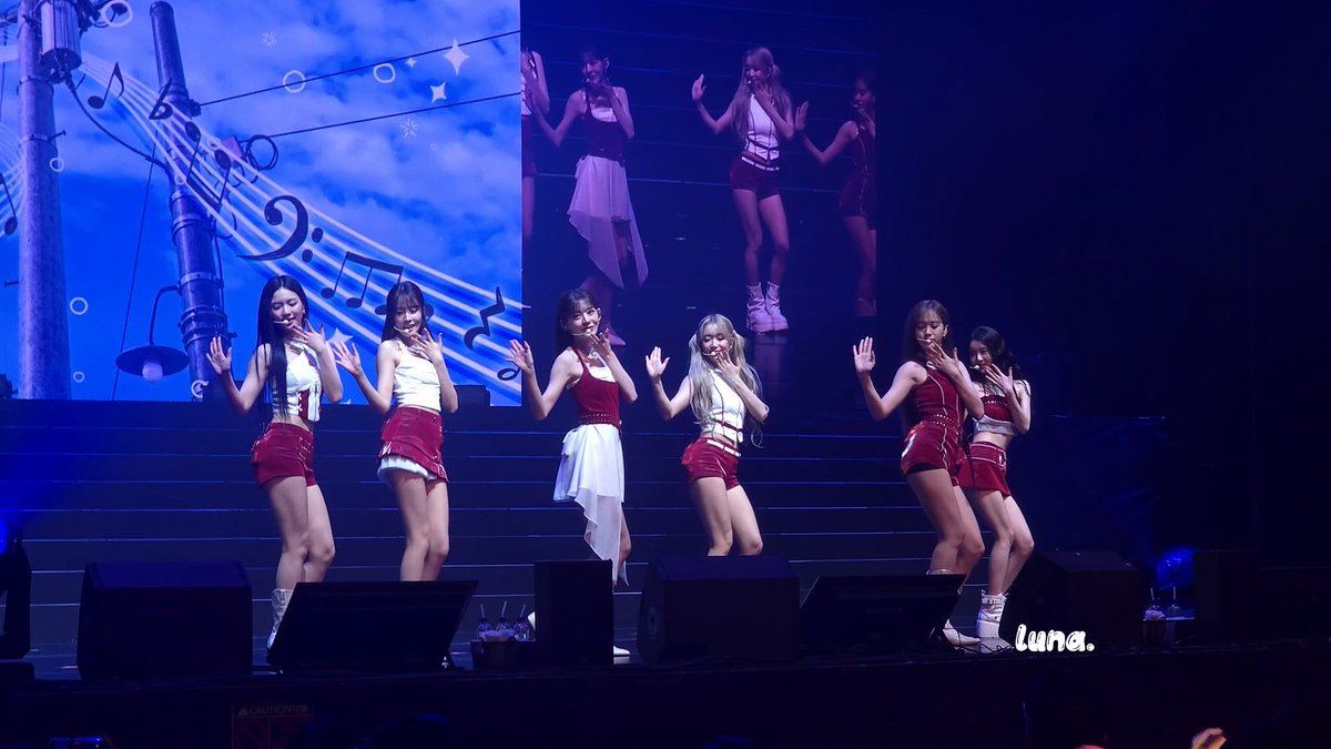 Six female K-pop performers in coordinating white and red stage outfits perform choreography on a tiered stage with a large screen displaying musical notes and a blue sky in the background; a 'luna.' watermark appears in the corner.