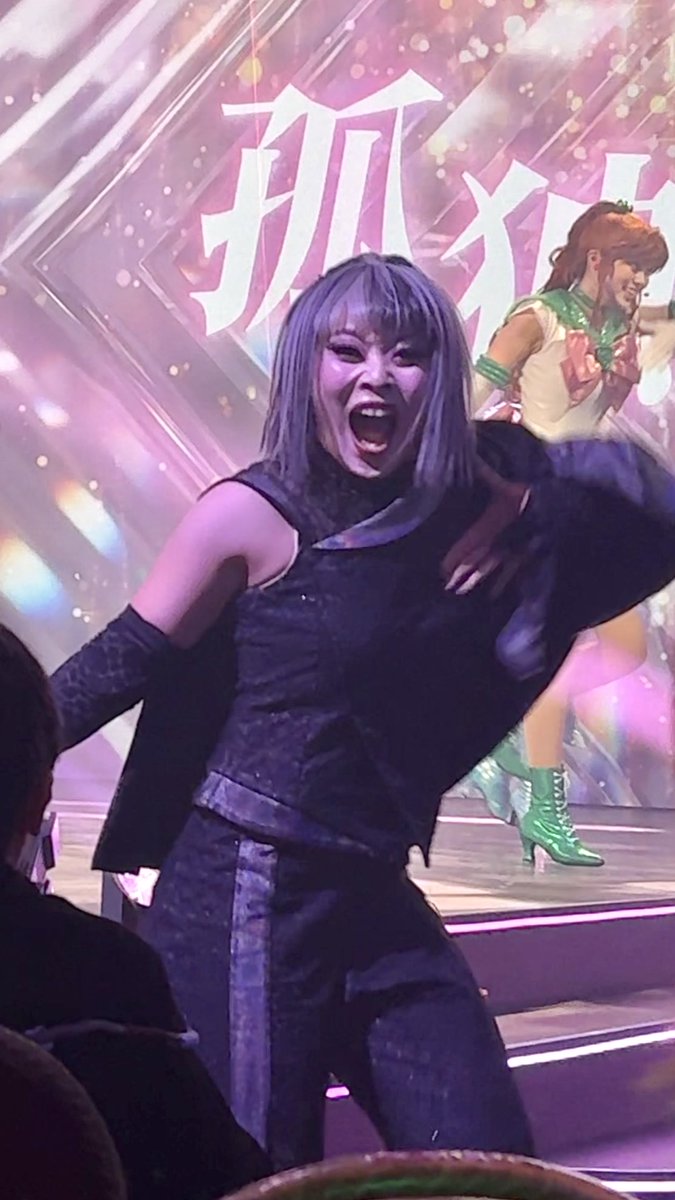 A performer on stage with a purple-streaked bob wig, mouth open mid-performance, wearing a dark sleeveless outfit in front of a backdrop featuring Japanese kanji characters.