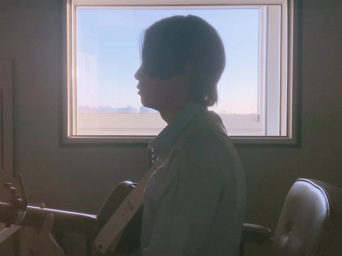 A silhouette of a young person with short hair playing an acoustic guitar in profile, backlit by bright sunlight streaming through a large window.