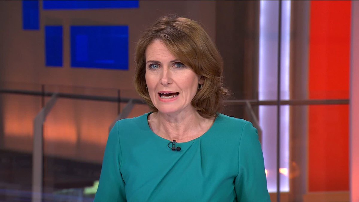 A female news presenter with shoulder-length brown hair, wearing a teal dress and a clip-on microphone, speaks directly to the camera in a television studio with blue and orange lighting elements in the background. The broadcast covers the breaking news of Amnesty International UK chair Sandy Ruthven's departure from the Green Party over their "Zionism is racism" motion.