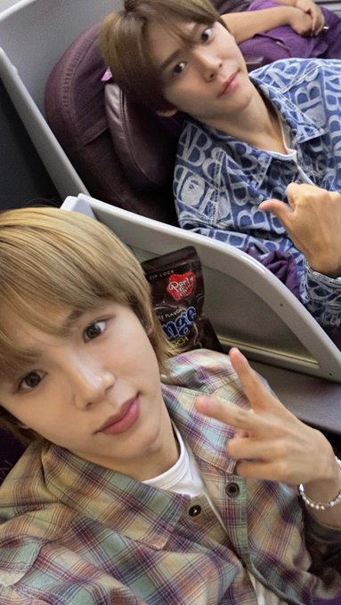 Two young men take a selfie on an airplane; one with blonde hair in a plaid shirt makes a peace sign in the foreground, while another with brown hair in a blue patterned shirt makes a hand gesture behind him, with bags of Party Mix snacks visible in the seatback pocket.