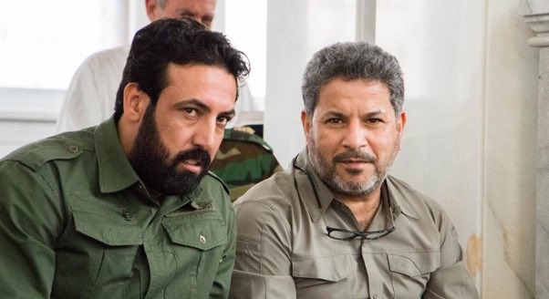 Two men sit closely together, with the younger bearded man on the left wearing a green military-style shirt and the older man on the right in a tan shirt with glasses hanging from it. The image relates to their discussion as it likely depicts Dr. Jaafar Dhia Jaafar (right) alongside his associate, reflecting the post's reference to key figures in Iraq's nuclear program.