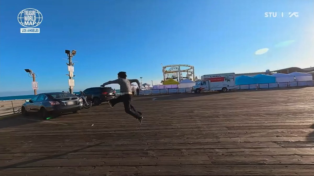 A person leaps joyfully in mid-air on a wooden boardwalk by the ocean in Los Angeles, with the 'TREASURE WORLD MAP' and 'STUDIO K' logos visible in the top corners.