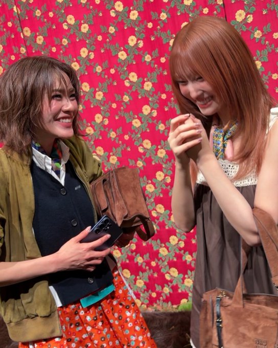 Two women smiling and laughing together against a red floral wallpaper background, with one holding a phone and brown bag while the other wears a floral scarf.