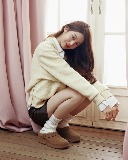 A young woman crouches by a window with pink curtains, wearing a cream sweater, white collared shirt, brown shorts, white socks, and tan suede boots.