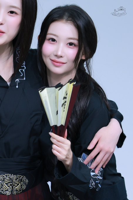 A young woman with long dark hair in a black traditional-style outfit poses holding a decorative folding fan.