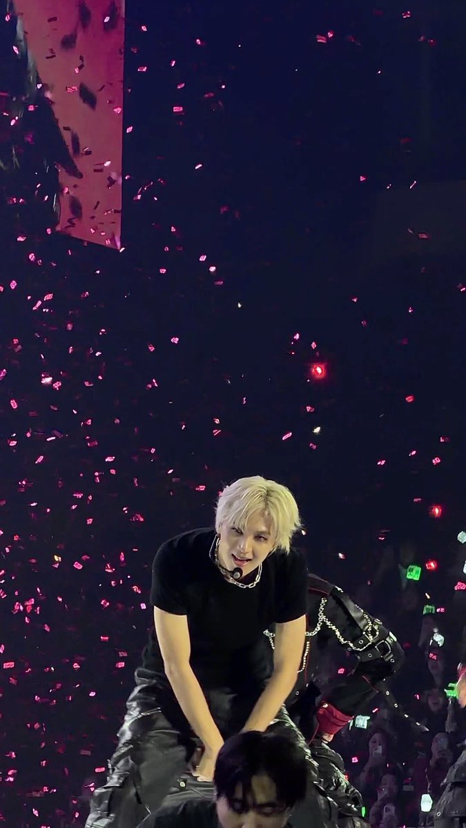 A blonde-haired performer in a black shirt performs on stage while kneeling on another performer's shoulders, surrounded by falling pink confetti and an energetic crowd with glowing light sticks.