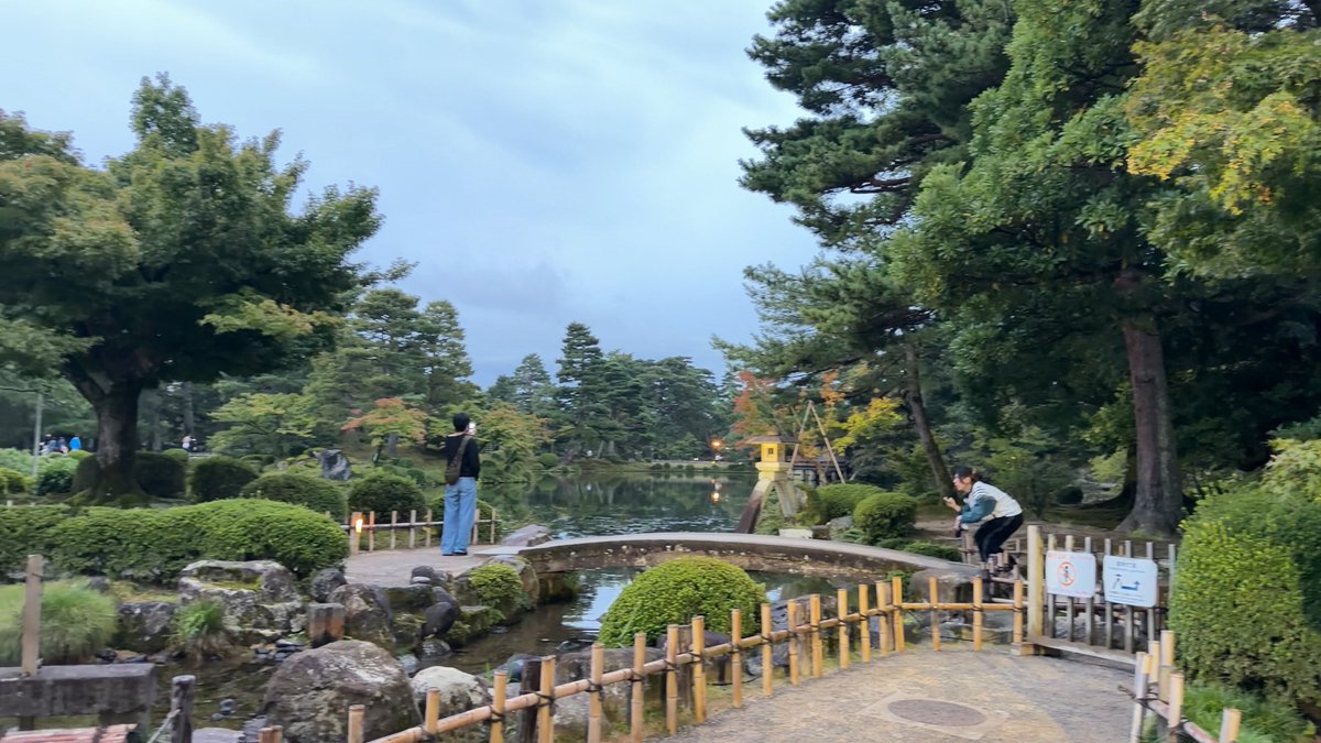 A tranquil Japanese garden scene with a pond, stone bridge, and manicured shrubs is disrupted by two men running through the frame, perfectly matching the caption about photobombing "running men" ruining the elegant atmosphere of Kenrokuen Garden.