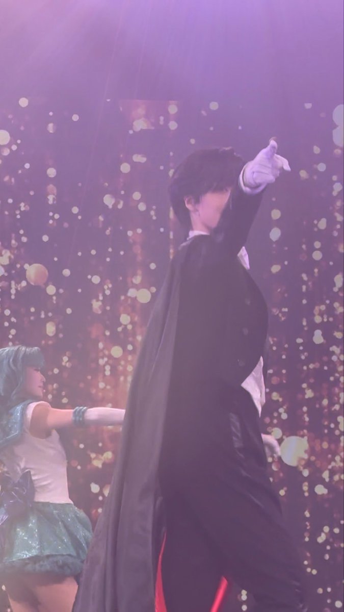 A performer in a black cape and white gloves points forward on a stage with sparkling lights, while a person in a blue costume stands to the left.