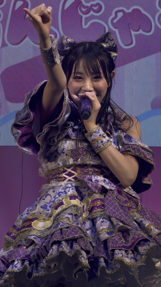 A young woman in an elaborate purple, white, and gold frilly dress performs on stage, holding a purple microphone and pointing upward, with a Hello Kitty pattern on the screen behind her.