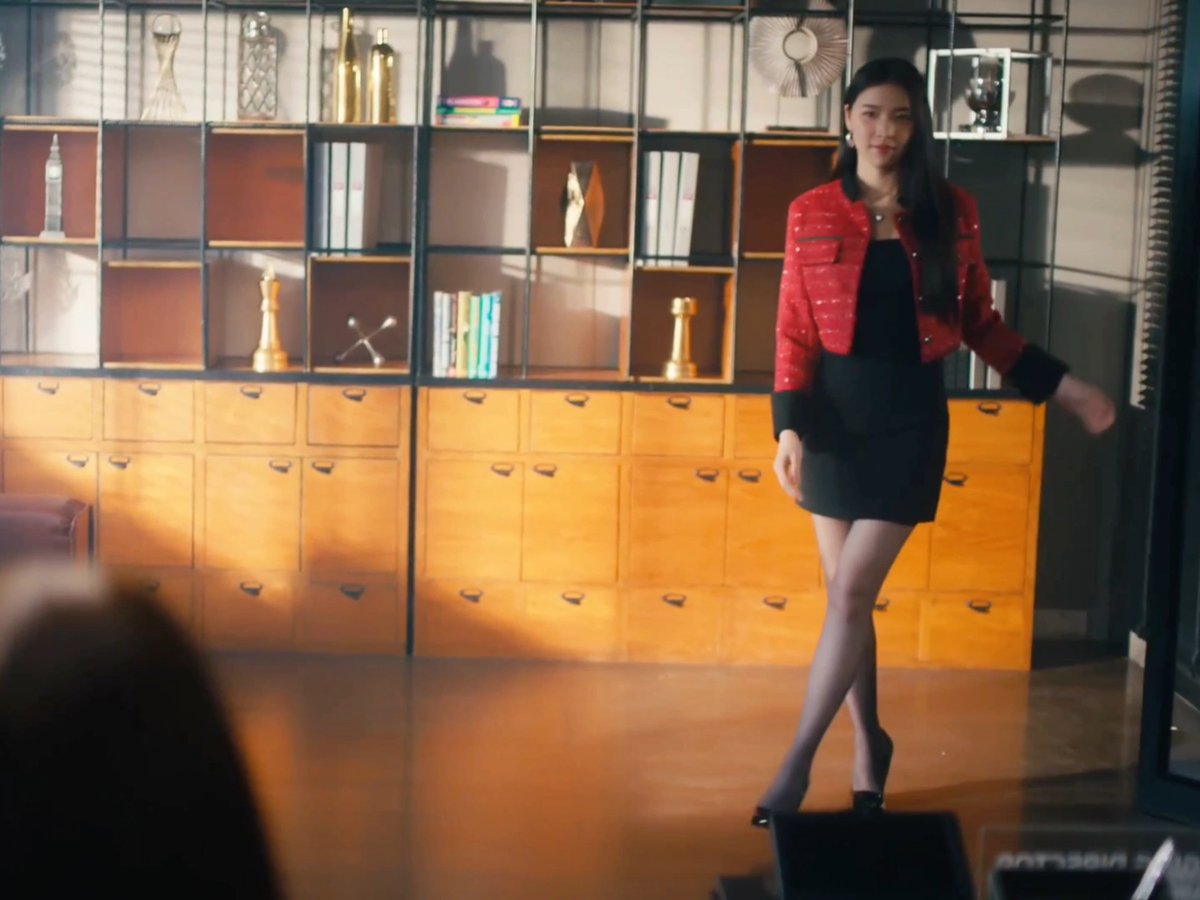 A woman with long dark hair wearing a red tweed jacket and black mini dress walks through an office featuring wooden shelving units with various decorative items.