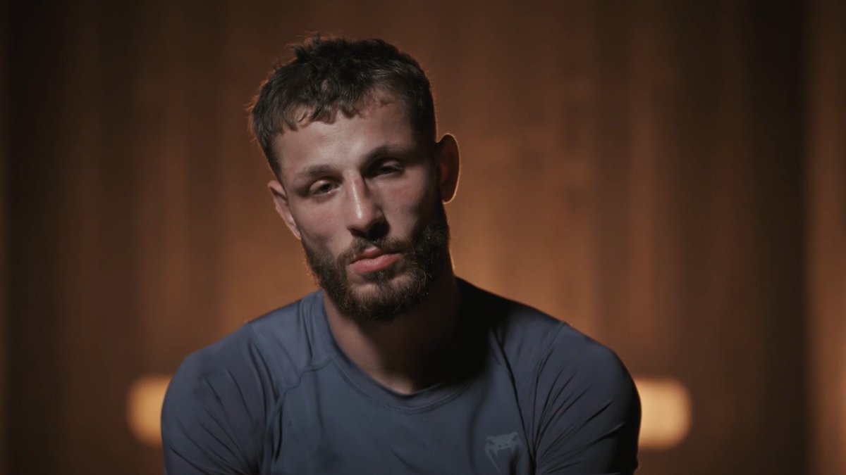 A bearded man with a serious, hardened expression stares directly at the camera, wearing a gray athletic shirt with a small logo on the chest. The moody lighting and intense gaze visually echo the post's reference to conflict, as his stern demeanor suggests the heated confrontation described between Joey and Mathis.