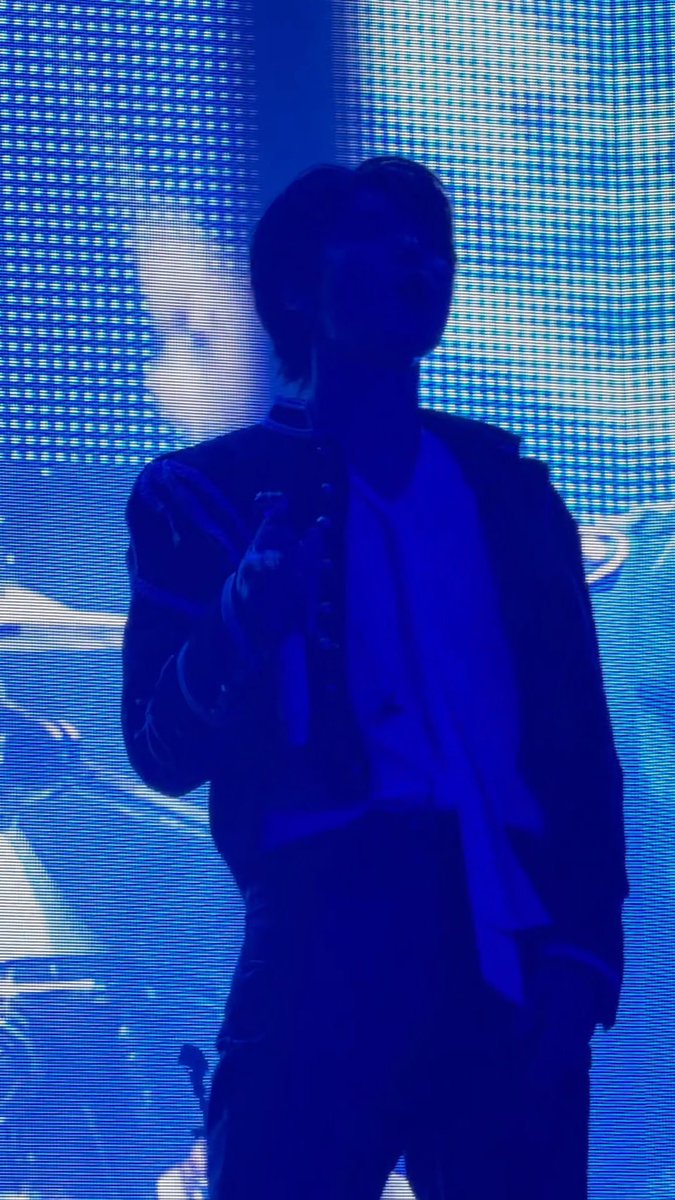 A performer on a stage bathed in blue lighting holds a microphone, silhouetted against a bright LED screen backdrop.