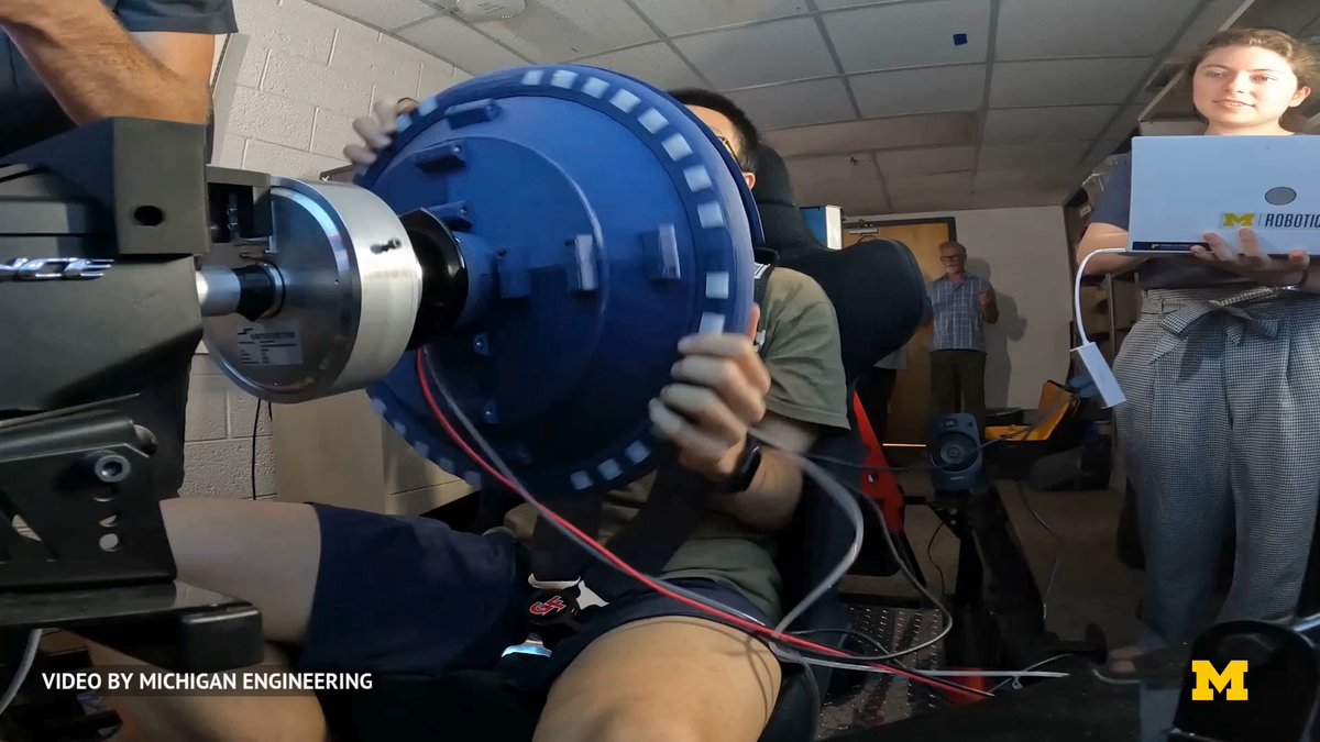 An engineering lab scene with people working on a large blue mechanical component attached to machinery, credited "VIDEO BY MICHIGAN ENGINEERING" with the University of Michigan logo.