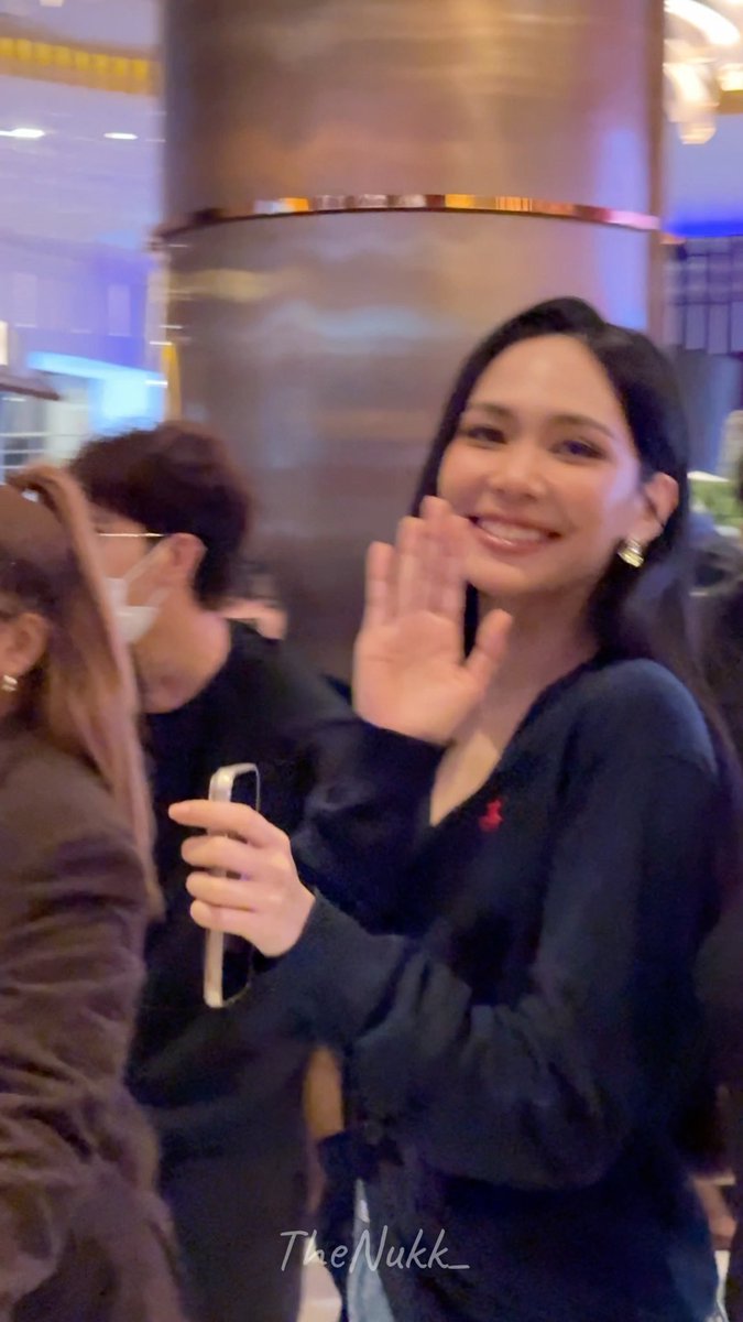 A smiling woman waves at the camera while holding her phone, wearing a black top and gold earrings in what appears to be a public venue. Watermarked 'TheNukk_'.
