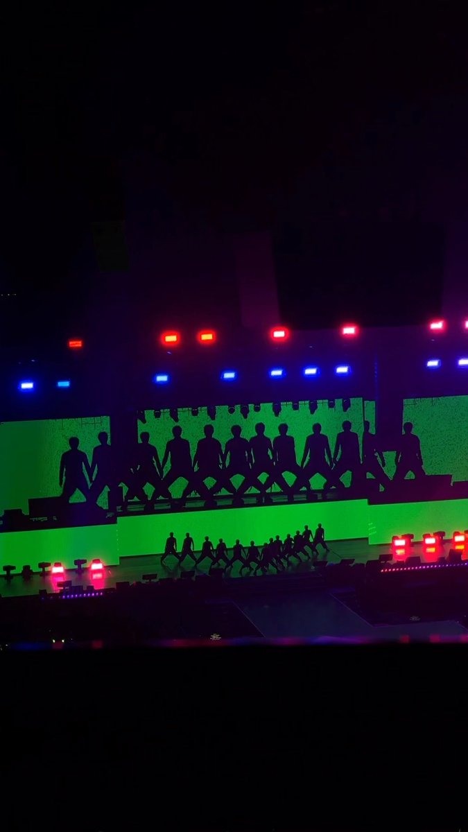 A concert stage with performers silhouetted against bright green lighting, with red and blue lights above.