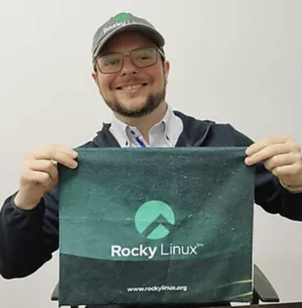 A smiling bearded man wearing glasses and a 'Rocky Linux' cap, holding up a green 'Rocky Linux' branded cloth or banner with the website www.rockylinux.org.