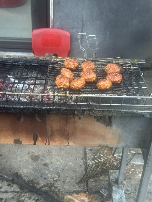 Photo of meatballs or small grilled patties cooking on a metal grate over a charcoal grill, with a red plastic chair and tongs visible in the background.