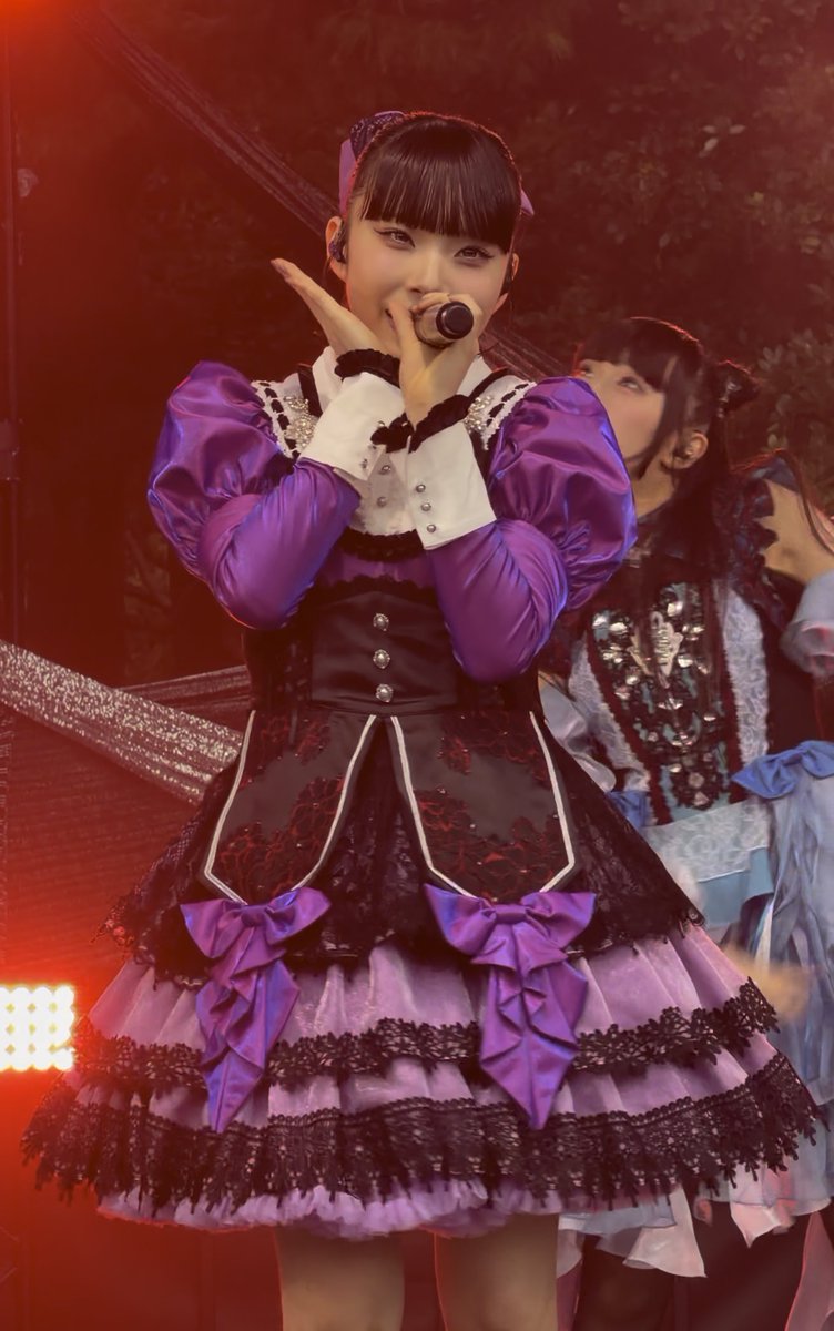 A Japanese pop idol performs on stage in a purple and black dress with ruffles, holding a microphone to her mouth with another performer visible behind her.