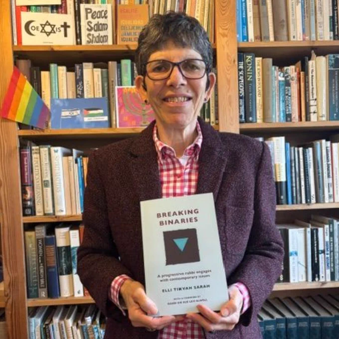 Colour photo of Elli Tikvah Sarah smiling at the camera whilst holding the book 'Beyond Binaries'
© Jess Wood