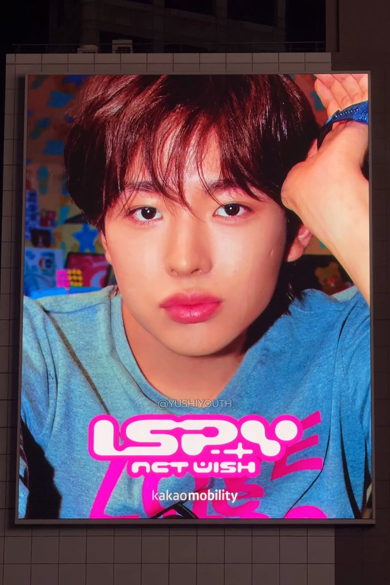 A large illuminated billboard displays a vibrant promotional image for NCT WISH's comeback, featuring a young man with dark hair in a blue shirt posing against a colorful background. The text "NCT WISH" and "kakaomobility" appears at the bottom, complementing the caption's excitement about seeing the SPY comeback advertisement prominently displayed on a large screen.