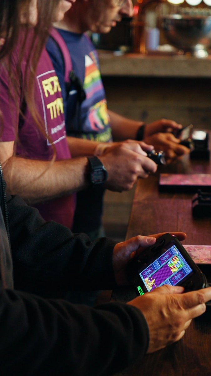 Close-up of people playing handheld gaming devices, including one showing a colorful match-3 style game on screen.