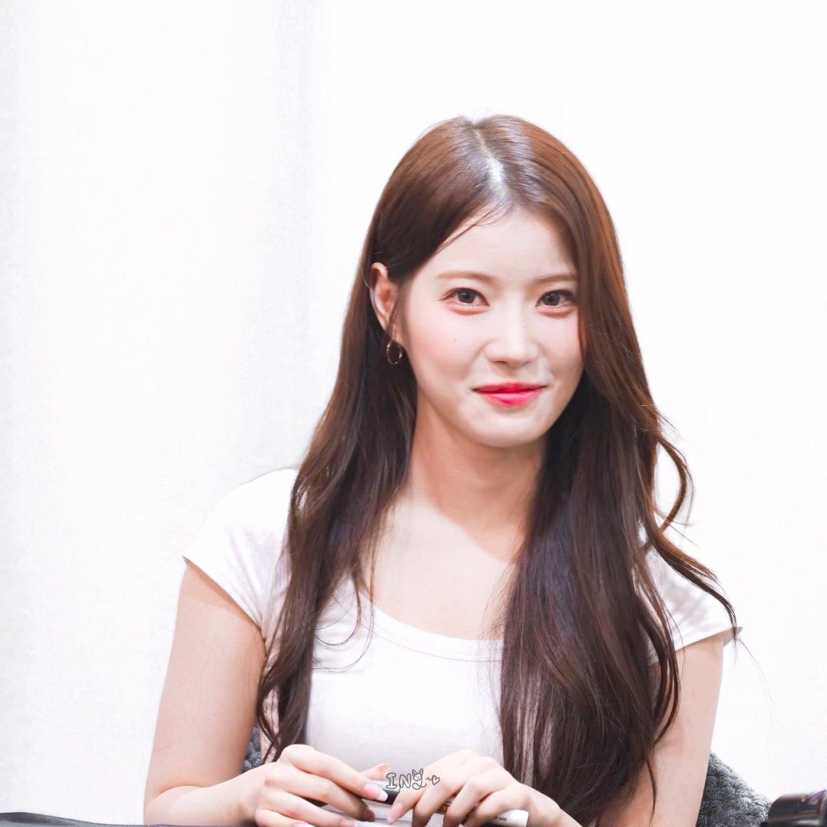 A smiling young woman with long brown hair, wearing a white t-shirt, poses with her hands clasped in front of her; the watermark "INU" appears at the bottom.