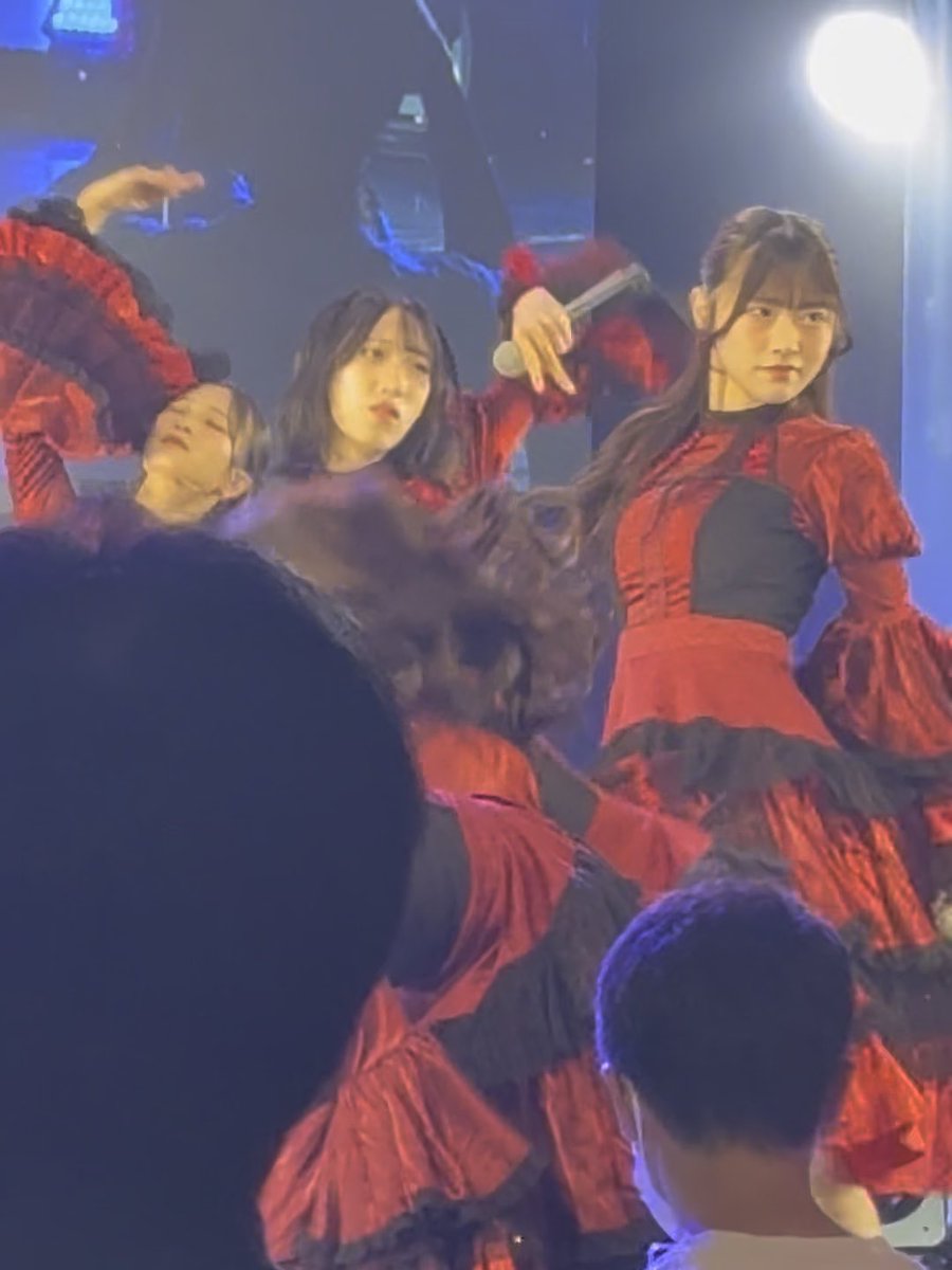 Several performers in dramatic red and black Victorian-style dresses perform on stage under blue and purple lighting, with audience members visible in the foreground.
