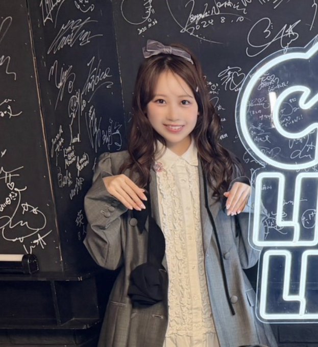 A young woman wearing a grey blazer over a cream ruffled top with a grey bow in her hair, posing in front of a black wall covered in signatures with a neon sign visible.