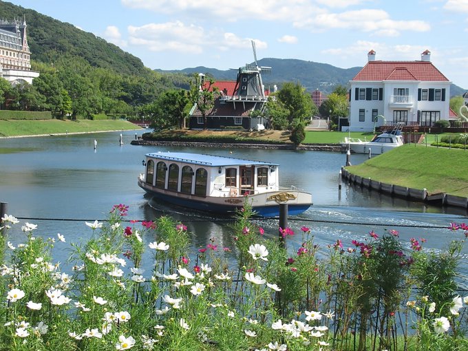 A scenic view of a boat cruising through calm waters with a traditional windmill, a Dutch-style white house with a red roof, and lush green hills in the background, framed by white and pink cosmos flowers in the foreground.