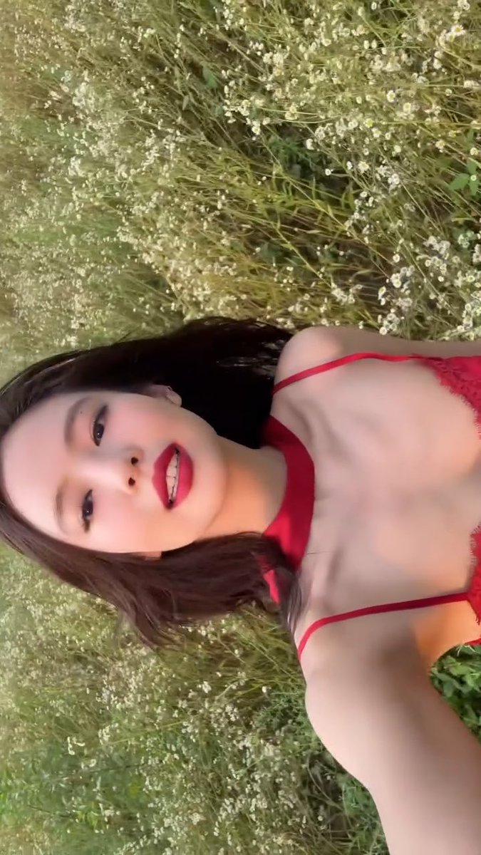 A selfie of a woman wearing a red top, taking a photo in a field of tall grass and small white flowers.