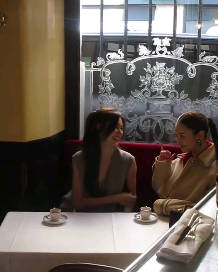 Two women sit across from each other at a white-clothed restaurant table with small coffee cups, smiling and conversing in front of an ornate etched glass window panel.