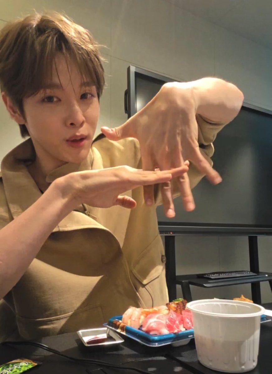 A young man with tousled brown hair, wearing a beige jacket, makes an expressive claw-like gesture with his hands toward the camera while seated at a table with sushi and a drink. The image appears to be a behind-the-scenes or vlog moment, possibly featuring Korean content creator Seongchan, whose expressive pose complements the post's playful tone about being "sucked in" to his immersive storytelling on a long flight.