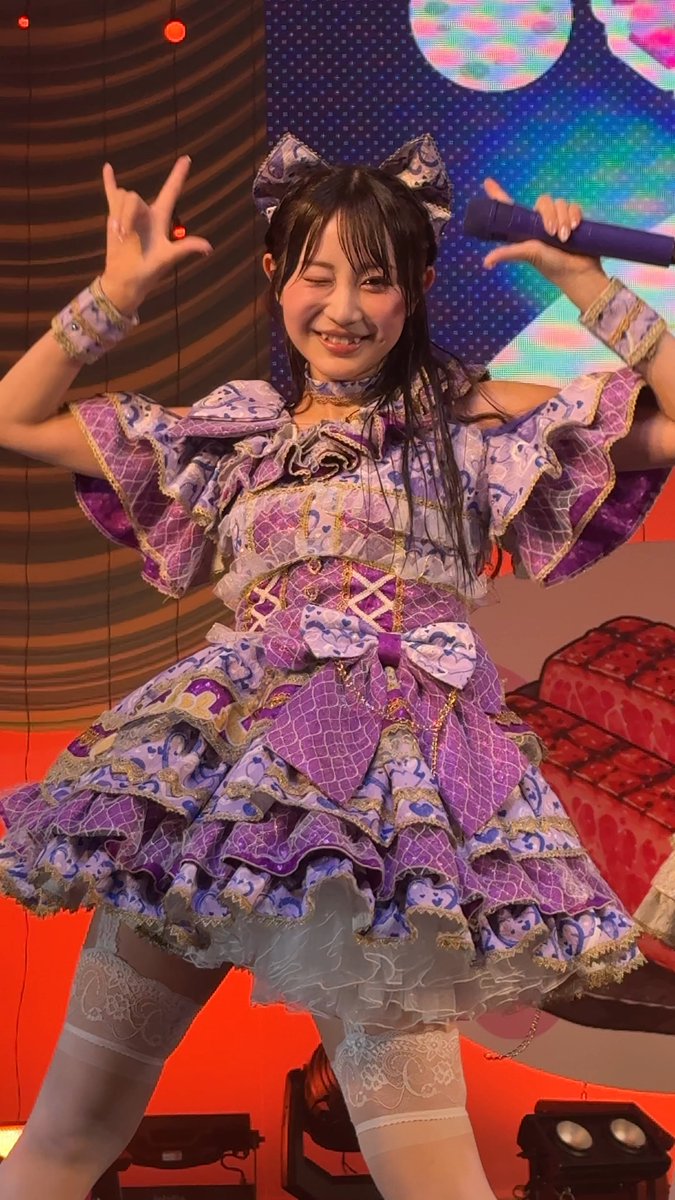 A Japanese idol performs on stage wearing an elaborate purple and gold frilly dress with matching headpiece, winking and making a rock hand gesture while holding a microphone.