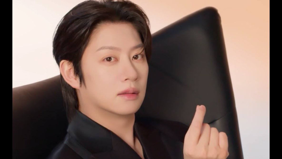 A close-up beauty-style photo of a young man in a black jacket, seated in a black chair and making a finger-heart gesture toward the camera.
