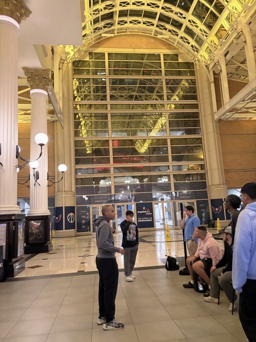 People line up inside an ornate building with a glass ceiling, waiting for "Comcast Center" doors to open, in anticipation of merchandise becoming available at 2 pm for a "PUREFLOW_IN_DC" event.