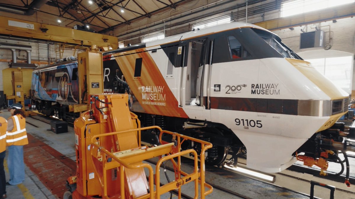 A modern white, orange, and black locomotive marked '91105' and labeled 'Railway Museum - 200 Years' sits in a maintenance depot alongside an orange cherry picker lift.