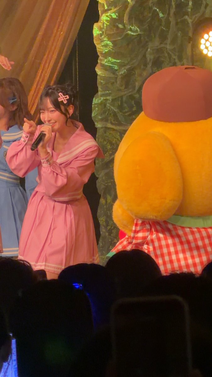 A young woman with dark hair wearing a pink sailor-style dress and a cross hair clip, performing on stage with a microphone next to a large yellow mascot character in a checkered apron.
