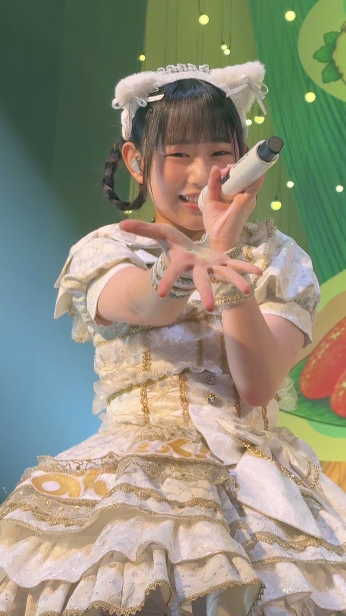 A young woman wearing a white and gold ruffled dress and cat-ear headband performs on stage, holding a microphone and extending her hand toward the camera.