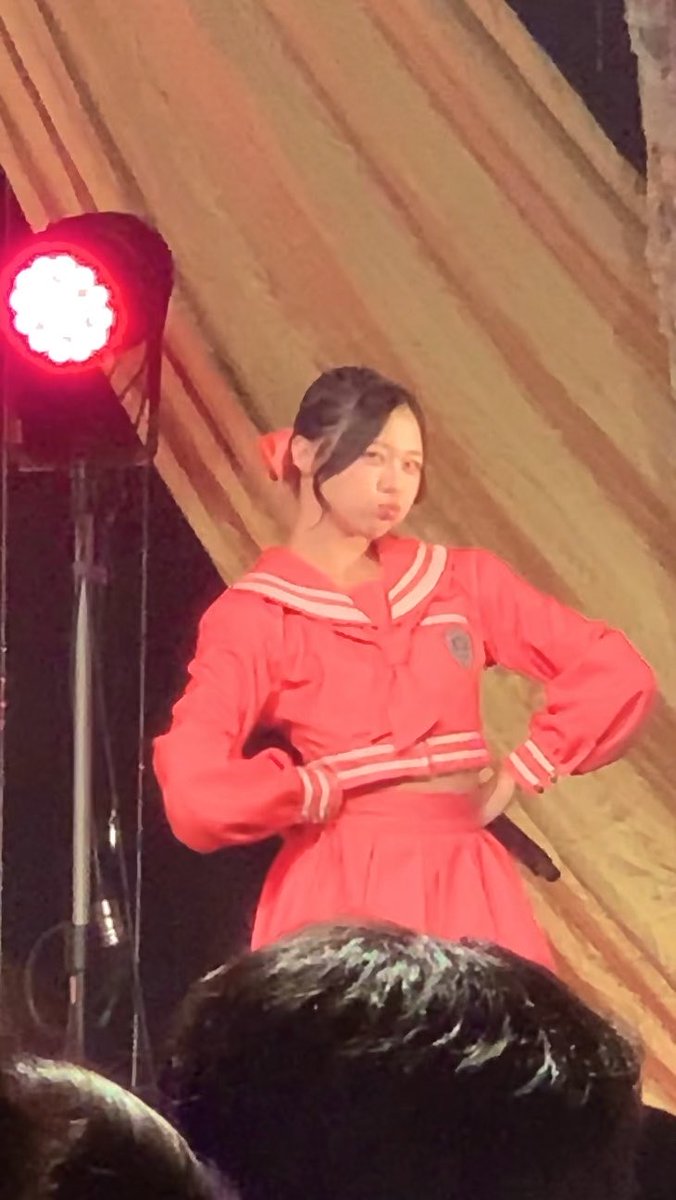 A young woman in a red sailor-style outfit performs on stage under a spotlight with a striped orange backdrop.