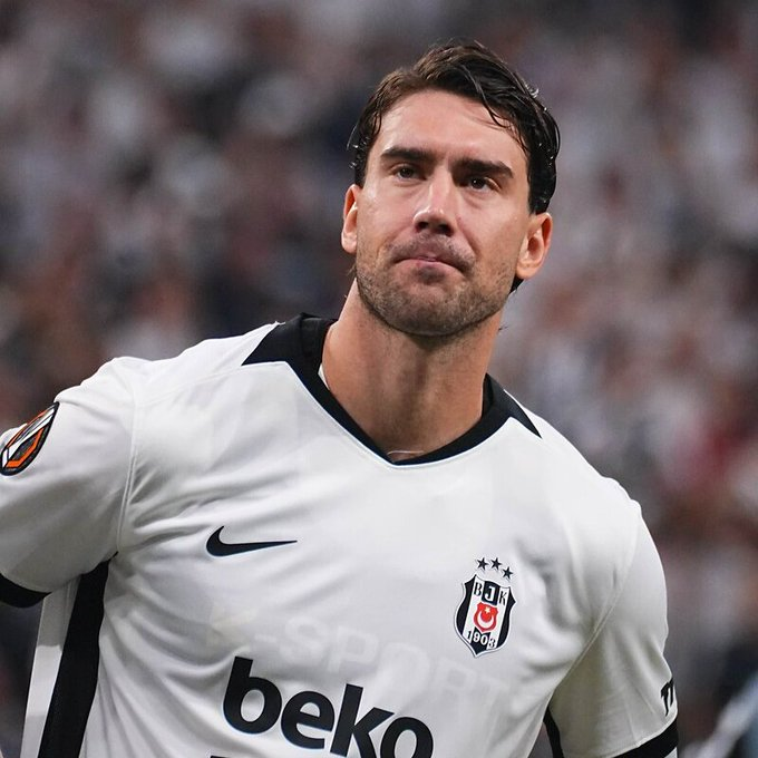 A close-up shot captures soccer player Dusan Vlahovic wearing a white Beşiktaş jersey featuring the Nike logo, club crest with three stars, and the "beko" sponsor text across the chest, along with a Europa League patch on the sleeve. The image aligns with the caption, showing Vlahovic beginning his tenure with Beşiktaş during a match.