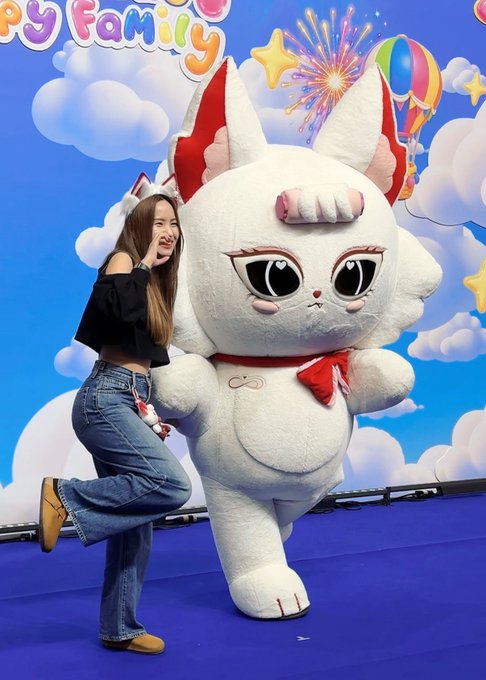 A young woman in a black top and blue jeans posing playfully next to a large white cat-like mascot character with red ears and a red bow at what looks like a "Happy Family" themed event.