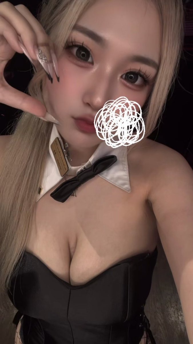 A close-up selfie shows a blonde woman with long nails wearing a black top and a white collar with a black bow, with a white scribble covering part of her face.