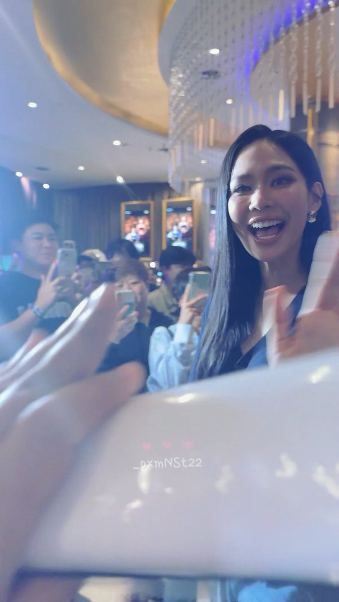 A smiling woman with long dark hair beams joyfully while extending her hand toward the camera, surrounded by excited fans holding up phones to capture the moment in an elegant venue with chandeliers. The watermark "_pxmNSt22" appears on a white surface in the foreground, matching the post's hashtags (#Bintsireethorn #puinoonbint) which suggest this is an enthusiastic fan meet-and-greet moment with the actress Bint Sireethorn, capturing the heartwarming connection she shares with her supporters.