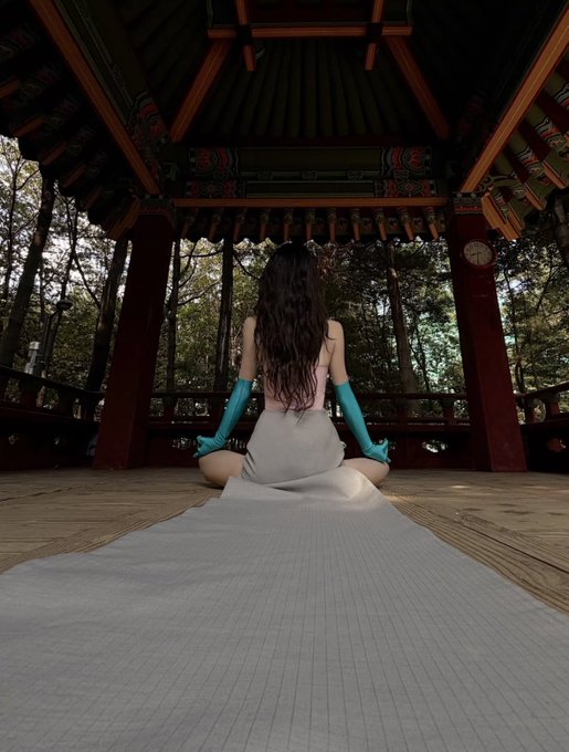 A woman with long wavy dark hair sitting on the floor of a traditional Korean pavilion (gazebo), wearing a white top, gray skirt with a long train, and turquoise gloves, viewed from behind.