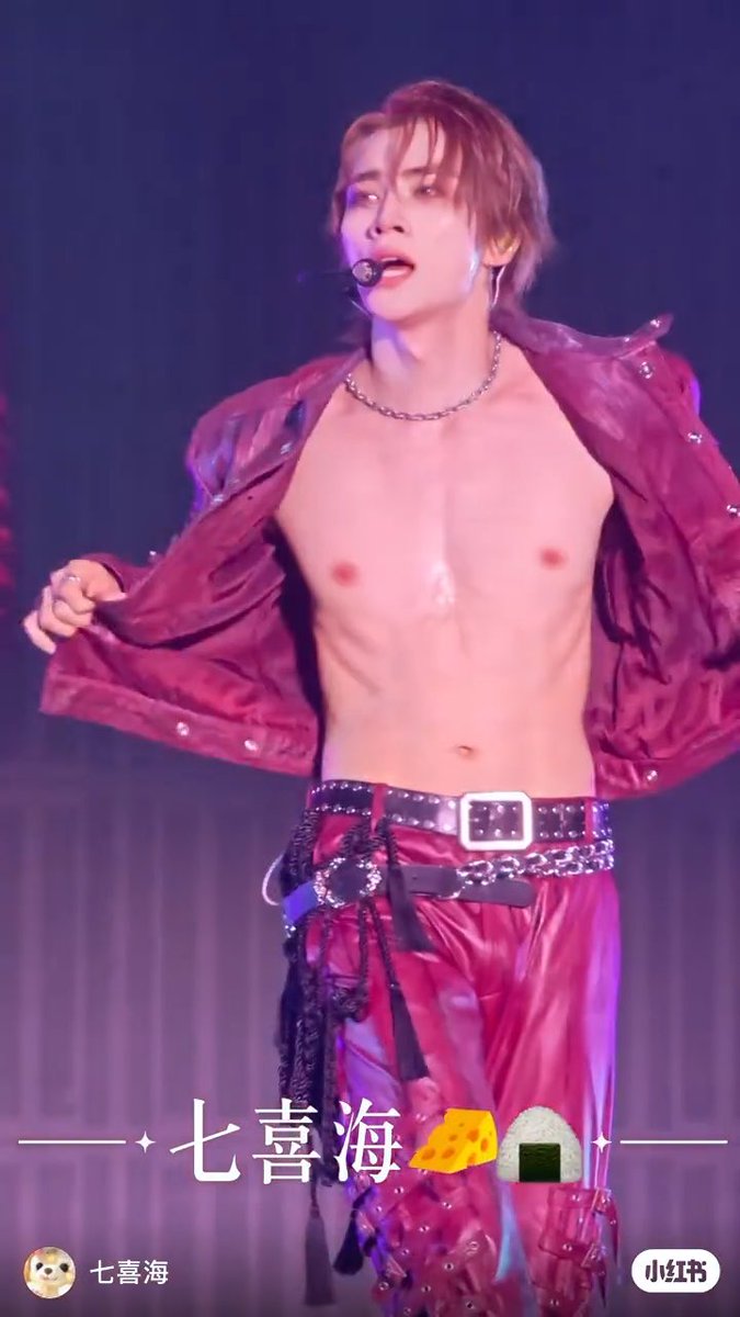 A male performer with reddish-brown hair performs on stage shirtless under an open red jacket, wearing a chain necklace and a headset microphone, with Chinese text '七喜海' overlaid.
