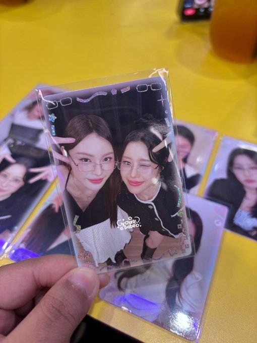 A hand holds a plastic-wrapped photocard showing two young women making peace signs and wearing pink glasses, with additional photocards scattered on a yellow table.