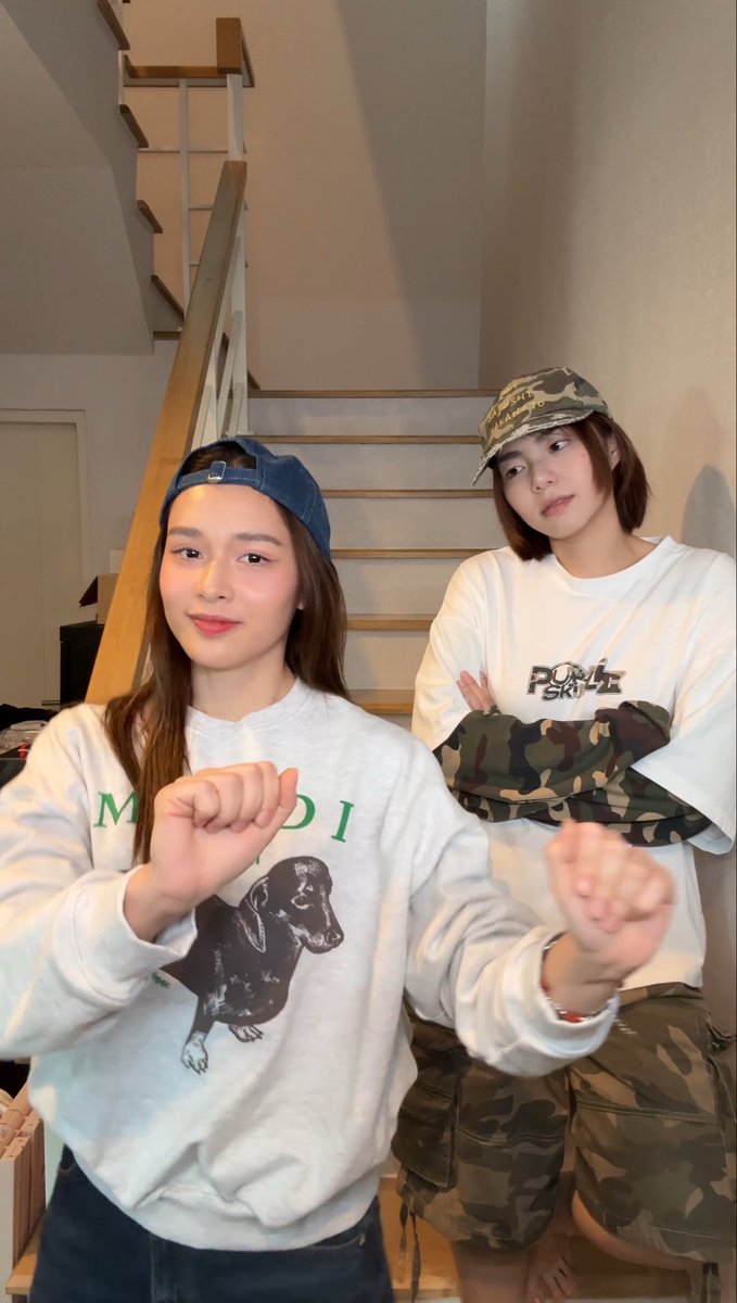 Two young women pose indoors near a staircase, one wearing a blue backwards cap and a white sweatshirt with a dachshund print, the other wearing a camouflage cap and a white 'POP SKATE' shirt with camo pants.